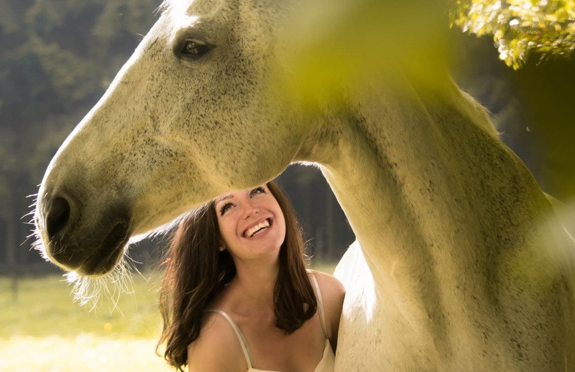 Eine lachende Frau neben einem Schimmel im Abendlicht